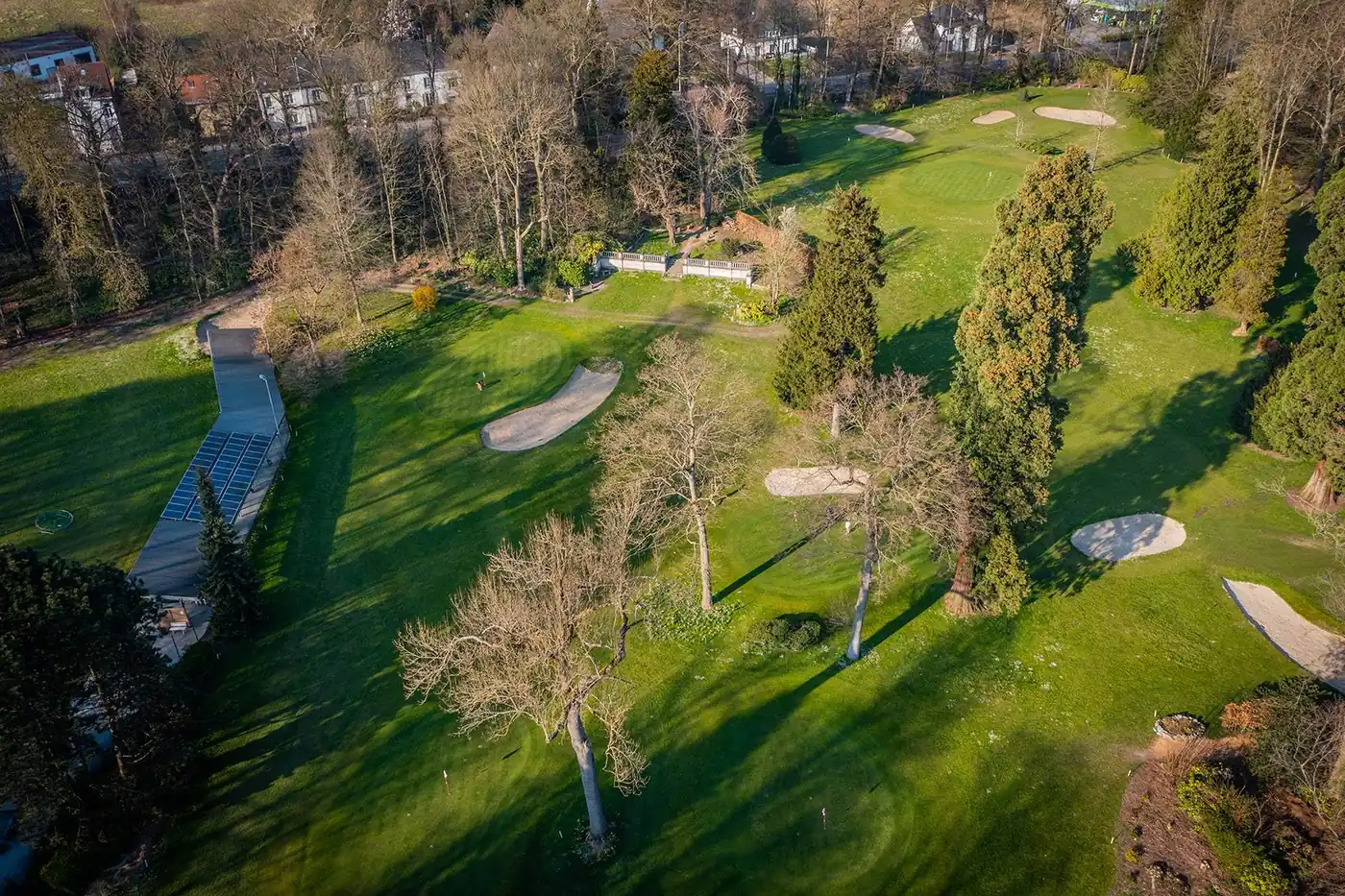 Luchtfoto van het parcours: greens, bunkers, oude bomen en de overdekte practice.